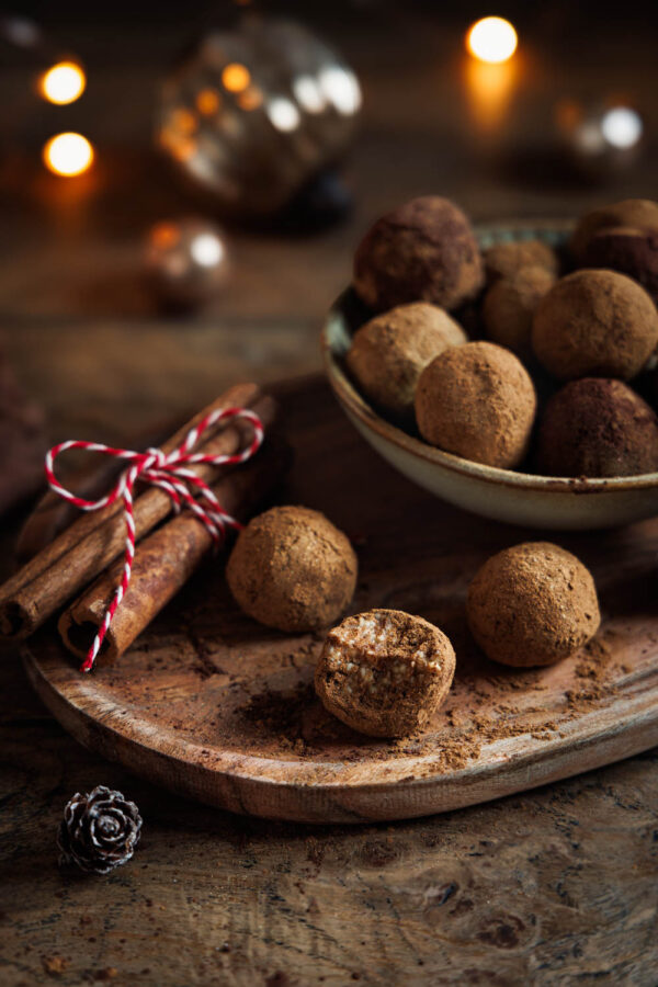 Selbstgemachte Spekulatius Pralinen auf rustikalem Holzbrett angerichtet, dekoriert mit Zimtstangen und weihnachtlicher Beleuchtung im Hintergrund – perfekt für die Festtage.