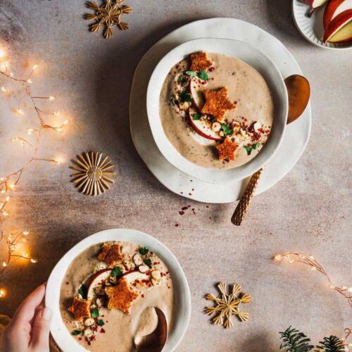 Schüssel mit cremiger Maronensuppe, dekoriert mit Apfelscheiben, gerösteten Nüssen, Petersilie und knusprigen Stern-Croutons. Weihnachtlich dekorierte Szenerie mit Lichterketten, Tannenzweigen und goldenen Schneeflocken.