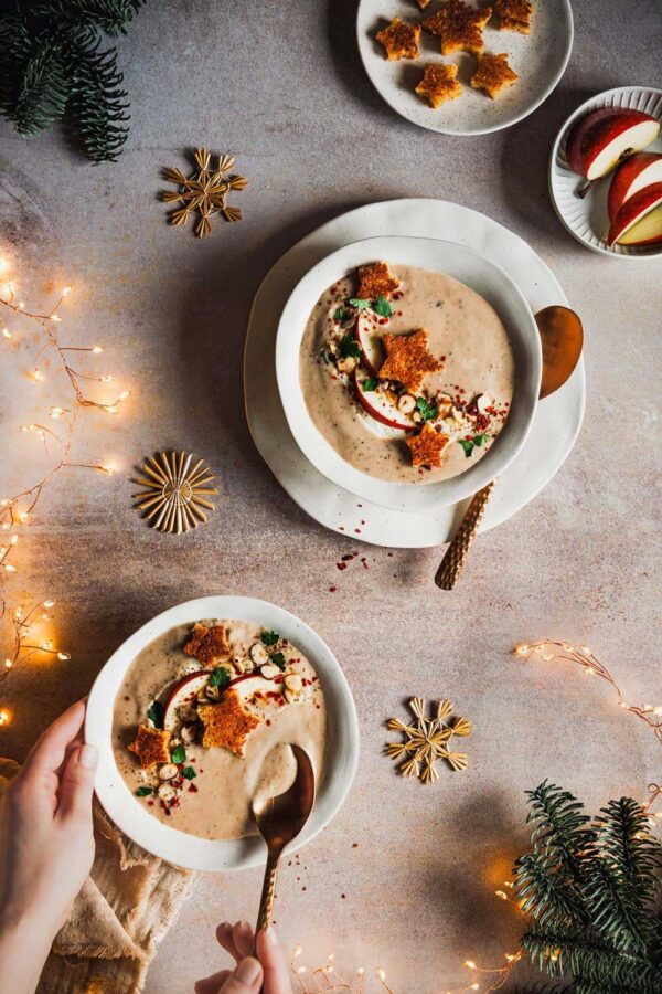 Schüssel mit cremiger Maronensuppe, dekoriert mit Apfelscheiben, gerösteten Nüssen, Petersilie und knusprigen Stern-Croutons. Weihnachtlich dekorierte Szenerie mit Lichterketten, Tannenzweigen und goldenen Schneeflocken.