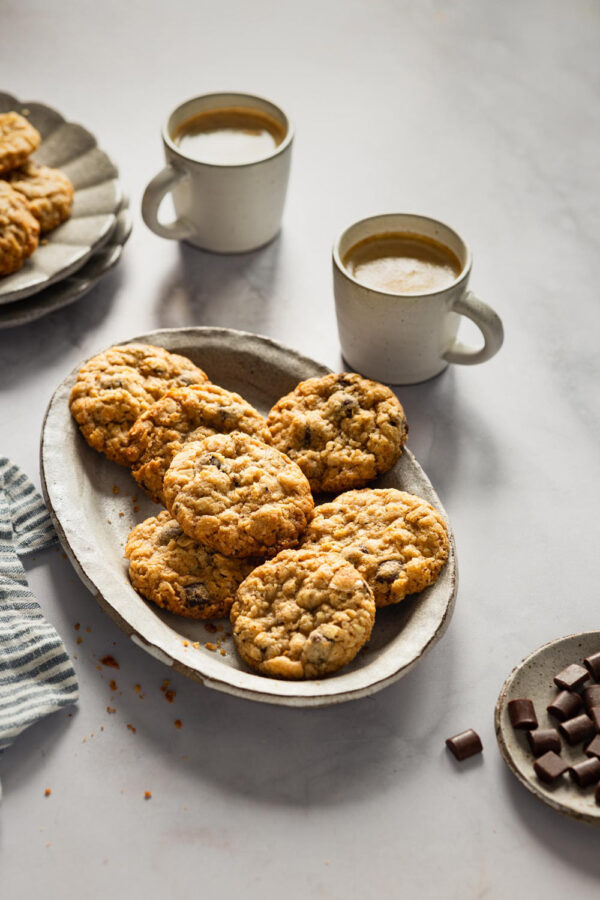 Eine rustikale Keramikplatte voller knuspriger schwedischer Hafercookies mit Sauerteig, umgeben von zwei Tassen dampfendem Kaffee in schlichten Keramikbechern. Im Vordergrund liegen Schokoladenstückchen.