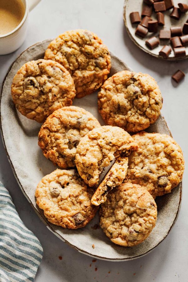 Goldbraune schwedische Hafercookies mit Sauerteig, angerichtet auf einer rustikalen Keramikplatte, umgeben von Zartbitterschokoladenstückchen und einer Tasse Kaffee – perfekt für eine gemütliche Auszeit.