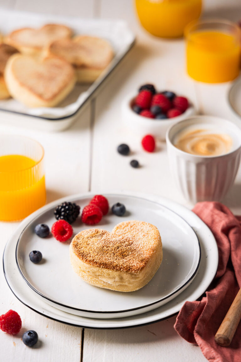 Herzförmiges Toastbrötchen auf einem Frühstücksteller, dekoriert mit frischen Beeren, Orangensaft und einem Cappuccino im Hintergrund. Perfektes Frühstücksarrangement mit liebevollen Details.