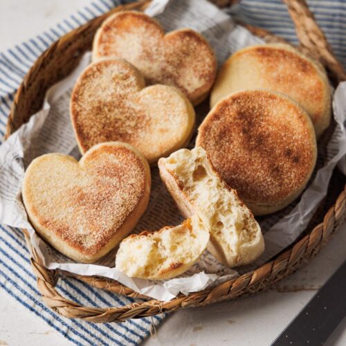 Herzförmige und runde Toastbrötchen in einem rustikalen Korb, serviert auf Papier und einem gestreiften Stofftuch. Ein aufgebrochenes Brötchen zeigt die weiche, luftige Krume.