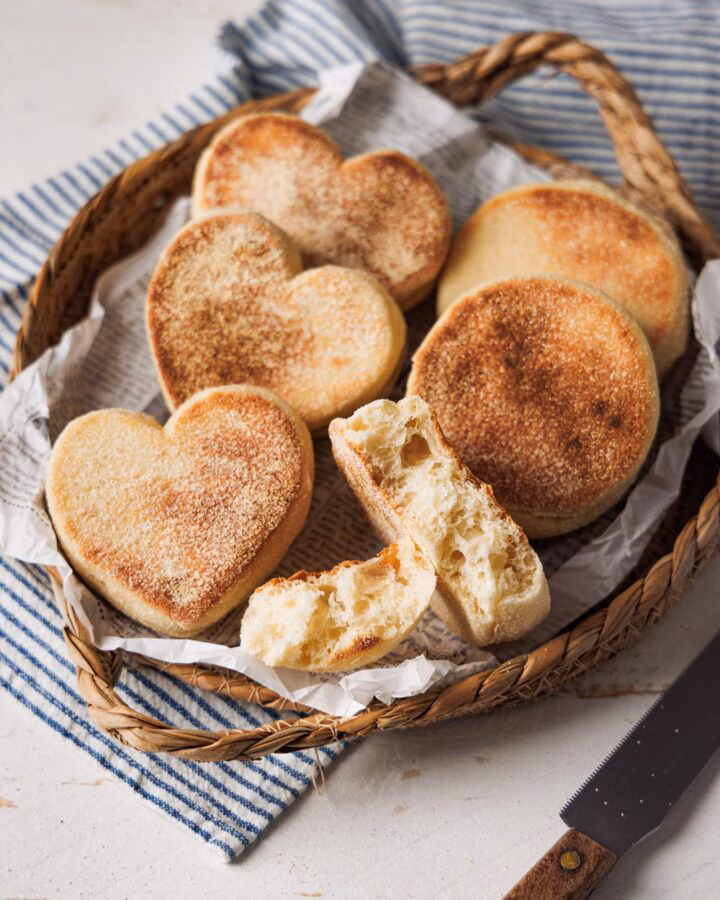 Herzförmige und runde Toastbrötchen in einem rustikalen Korb, serviert auf Papier und einem gestreiften Stofftuch. Ein aufgebrochenes Brötchen zeigt die weiche, luftige Krume.