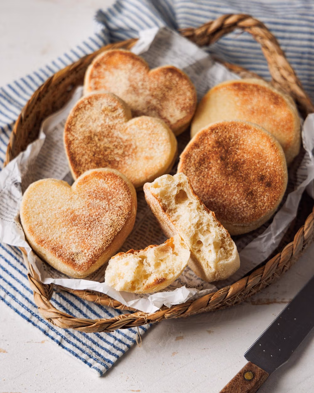 Herzförmige und runde Toastbrötchen in einem rustikalen Korb, serviert auf Papier und einem gestreiften Stofftuch. Ein aufgebrochenes Brötchen zeigt die weiche, luftige Krume.