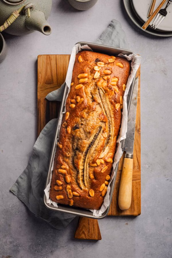 Ein Bananenbrot liegt in einer mit Backpapier ausgelegten Kastenform. Auf dem goldbraun gebackenen Teig sind eine halbierte Banane und Erdnüsse dekorativ angeordnet. Ein Messer mit Holzgriff liegt daneben.