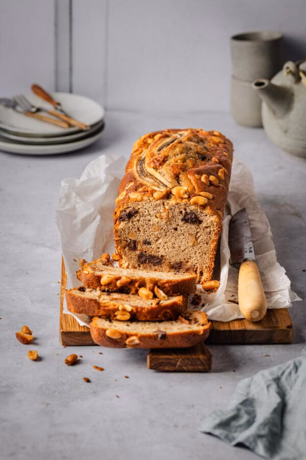 Das Bananenbrot ist auf einem Holzbrett angerichtet. Mehrere Scheiben wurden abgeschnitten und liegen im Vordergrund. Man sieht deutlich die Schokoladenstückchen im Teig sowie die gerösteten Erdnüsse obenauf.