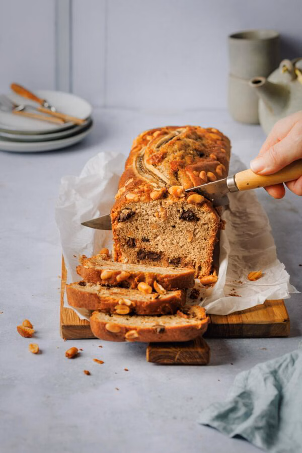Eine Nahaufnahme des Bananenbrots auf einem Holzbrett. Jemand schneidet mit einem Messer weitere Scheiben ab. Das Brot ist mit Erdnüssen und einer halbierten Banane verziert.