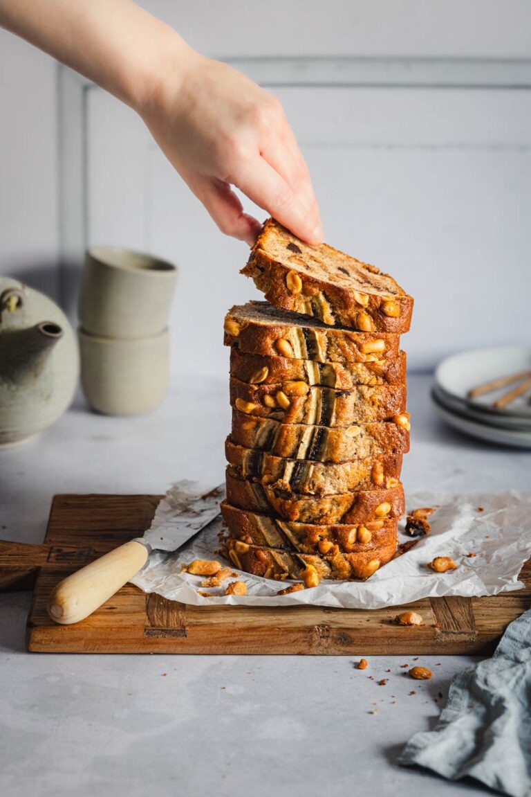 Mehrere Scheiben des Bananenbrots sind übereinandergestapelt. Eine Hand hebt die oberste Scheibe an. Auf dem Brot sind Erdnüsse und eine dekorative Bananenhälfte zu erkennen.