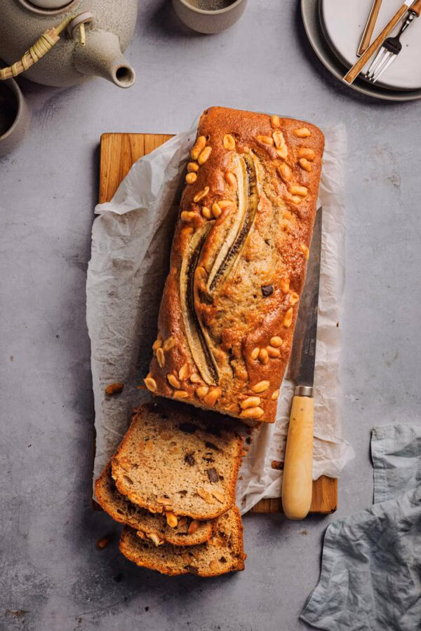 Ein frisch gebackenes Bananenbrot mit Schokoladenstückchen und Erdnüssen auf einer Holzunterlage. Einige Scheiben sind bereits angeschnitten, daneben liegt ein Messer. Im Hintergrund sind eine Teekanne und Teller zu sehen.