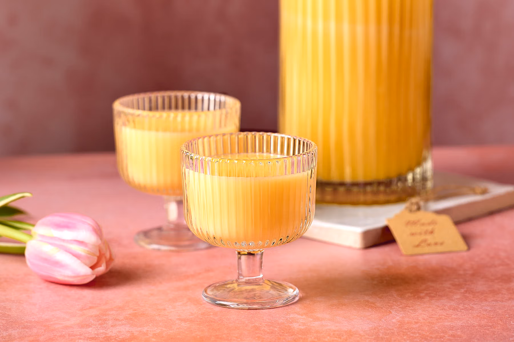 Eine Flasche mit gelbem Eierlikör und zwei gefüllte Gläser, dekoriert mit rosafarbenen Tulpen.