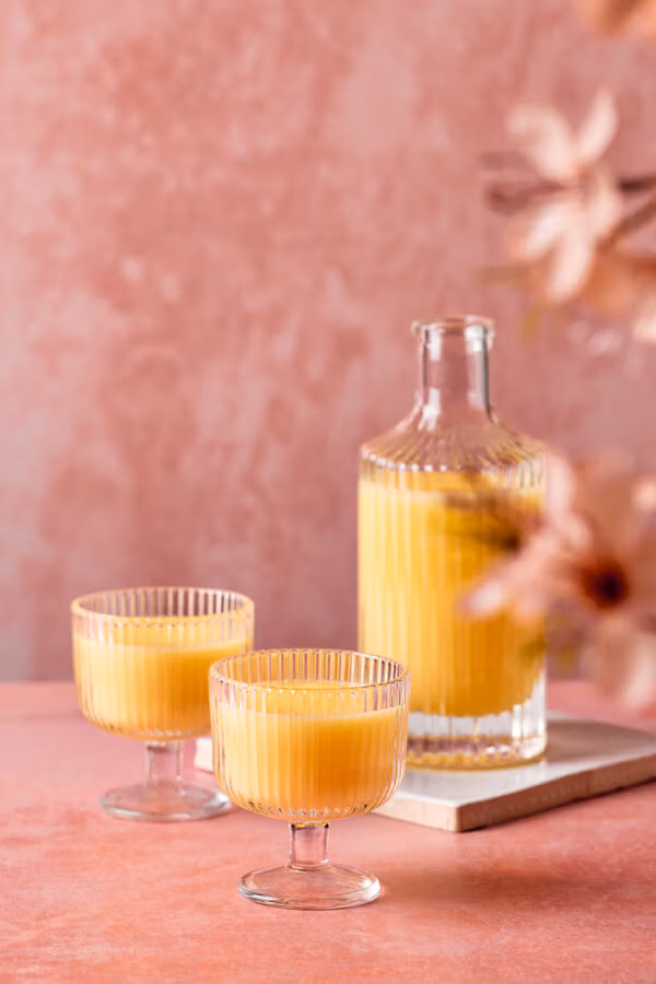 Zwei geriffelte Gläser mit cremigem Eierlikör vor einer passenden Karaffe. Der rosafarbene Hintergrund und einzelne Blüten im Vordergrund verleihen dem Bild eine frühlingshafte Stimmung.