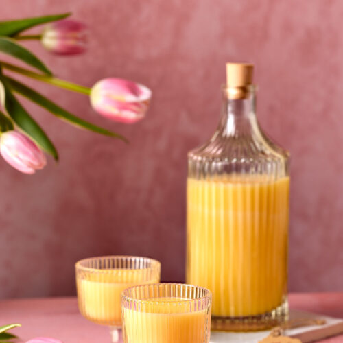 Eine Flasche mit gelbem Eierlikör und zwei gefüllte Gläser, dekoriert mit rosafarbenen Tulpen. An der Flasche hängt ein kleines Etikett, auf dem ‚Eierlikör‘ steht.