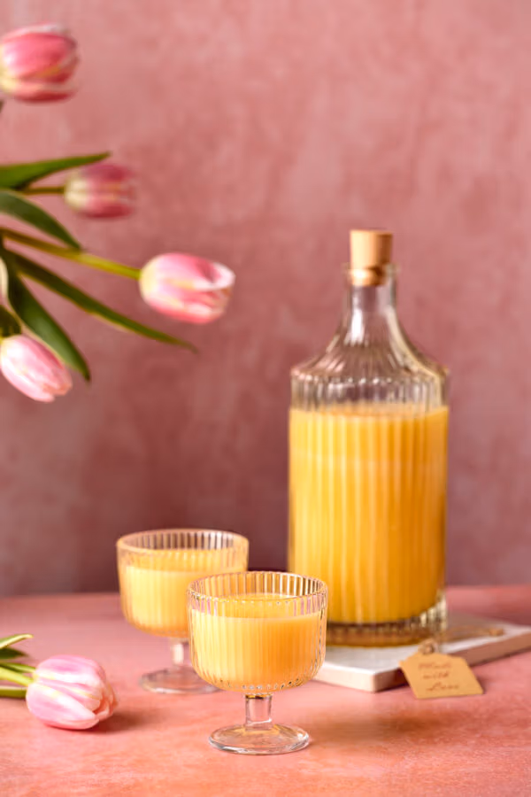 Eine Flasche mit gelbem Eierlikör und zwei gefüllte Gläser, dekoriert mit rosafarbenen Tulpen. An der Flasche hängt ein kleines Etikett, auf dem ‚Eierlikör‘ steht.