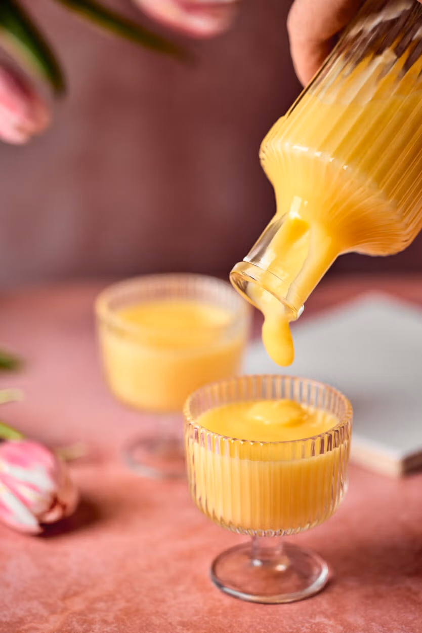 Nahaufnahme beim Einschenken des cremigen Eierlikörs aus einer geriffelten Glasflasche in ein passendes Glas. Im Hintergrund liegen rosafarbene Tulpen.