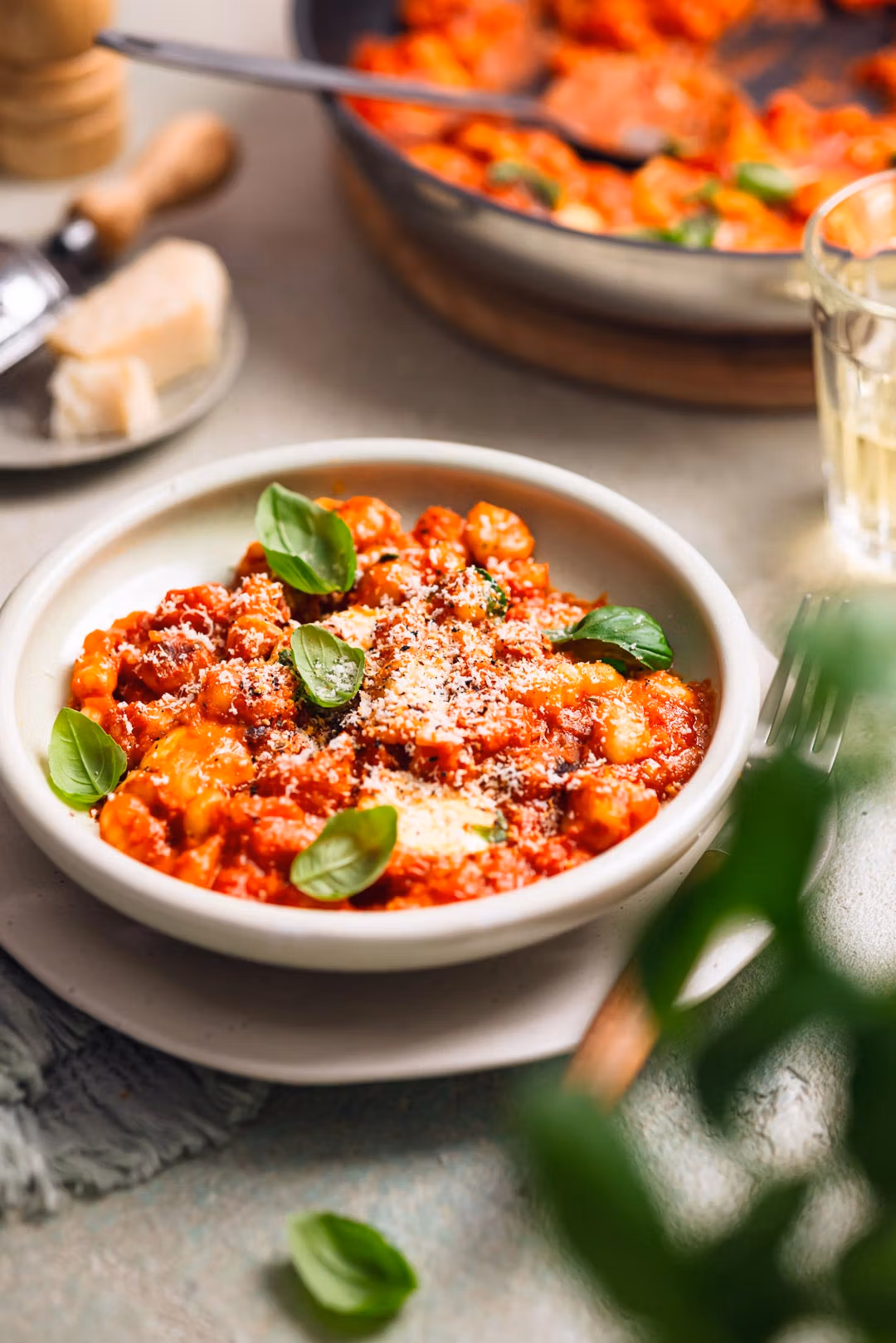 Eine Schüssel mit Gnocchi in Tomatensauce, bestreut mit Parmesan und garniert mit Basilikum. Im Hintergrund sind eine Pfanne mit derselben Speise und ein Glas zu erkennen.