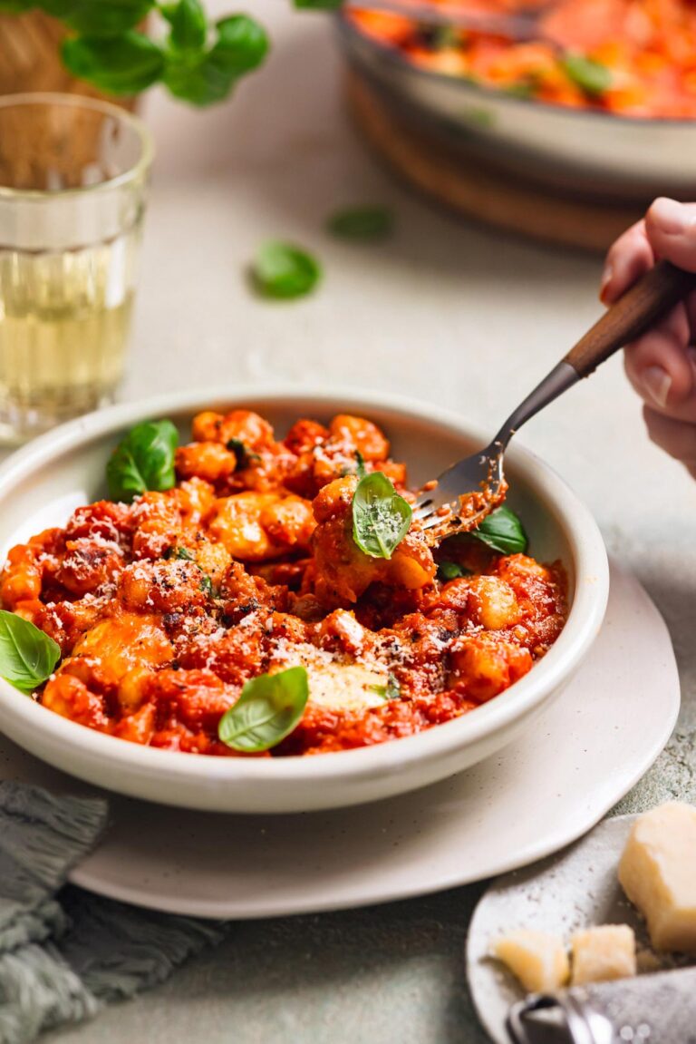 Nahaufnahme von Gnocchi in Tomatensauce, bedeckt mit Käse und frischen Basilikumblättern. Eine Hand sticht mit einer Gabel ein Stück Gnocchi auf.