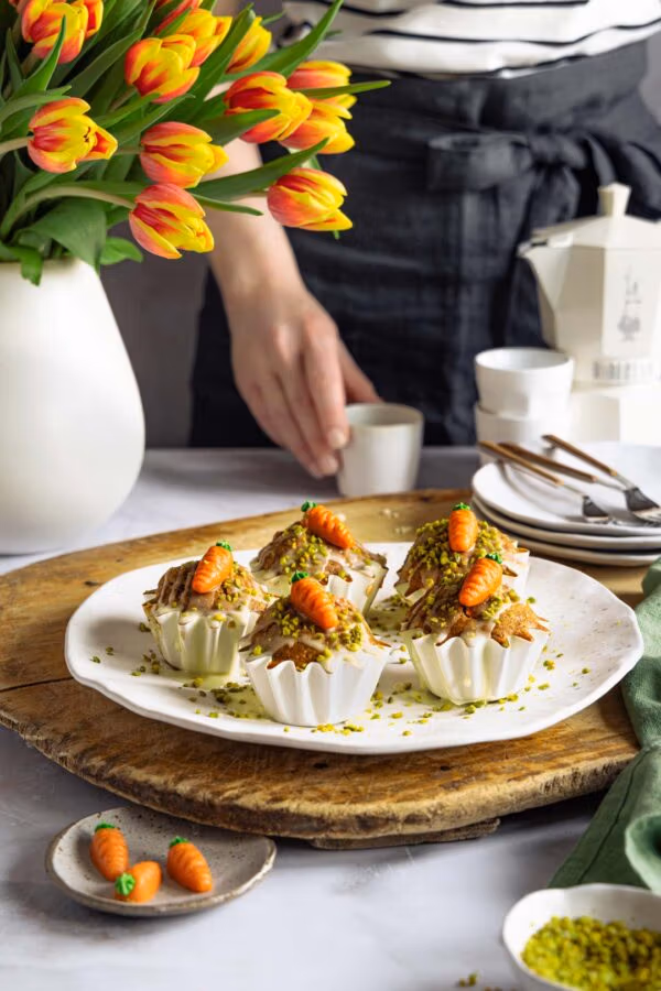 Mehrere Rüblimuffins auf einem weißen Teller, garniert mit Pistazien und Marzipankarotten. Dahinter steht eine Person in dunkler Schürze, neben einer weißen Espressokanne und einem Strauß Tulpen.