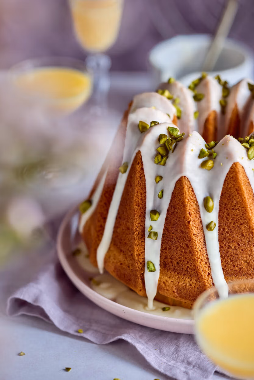 Ein goldbraun gebackener Eierlikörkuchen in Gugelhupfform. Der Kuchen ist mit einer hellen Eierlikör-Glasur überzogen und mit gehackten Pistazien bestreut. Daneben stehen kleine Gläser mit Eierlikör.