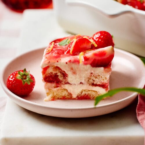 Eine Portion Erdbeer-Tiramisu auf einem Teller mit einer Gabel, einem Schälchen mit frischen Erdbeeren und Minzblättchen im Vordergrund und eine Keramikform mit dem Tiramisu im Hintergrund.