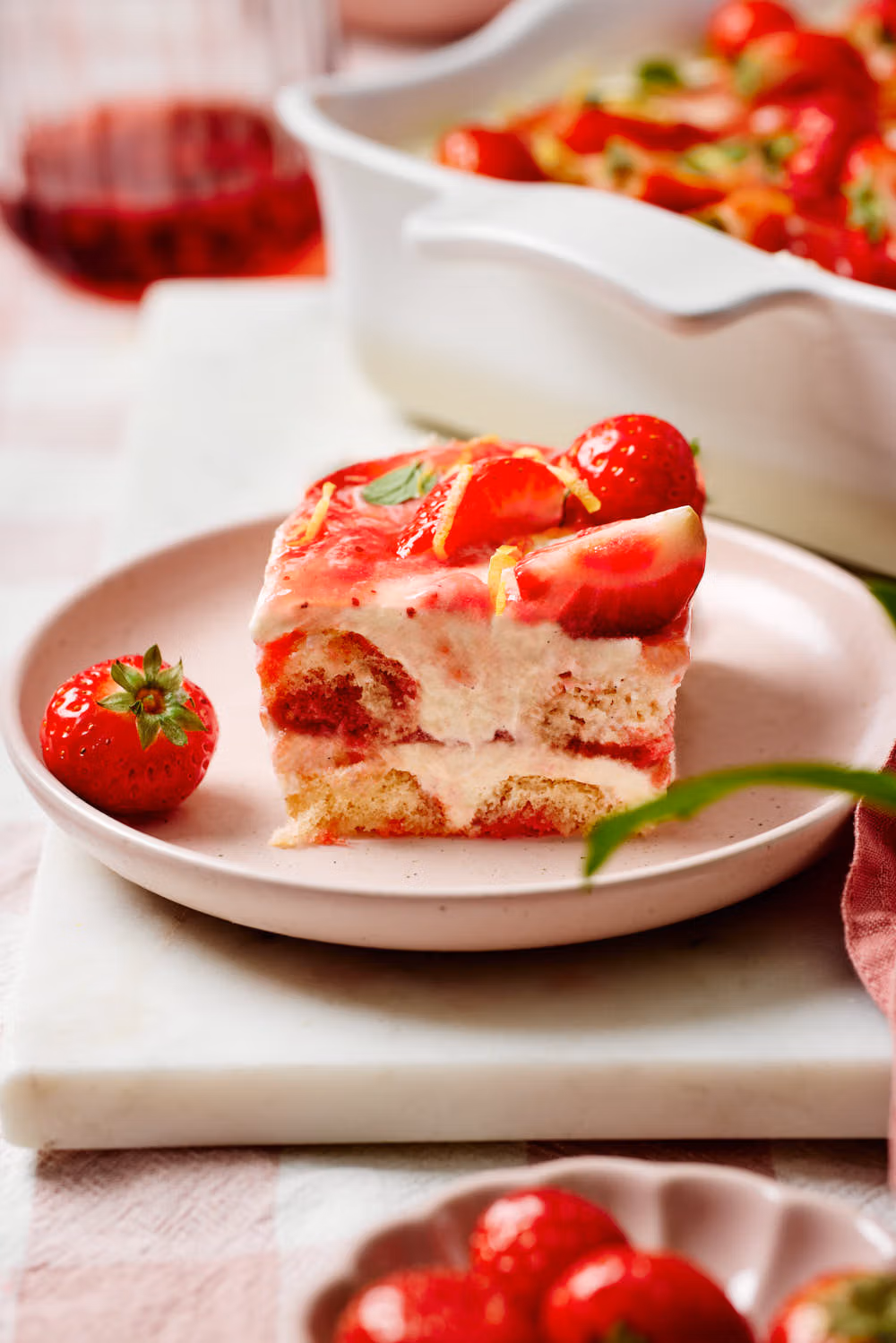 Eine Portion Erdbeer-Tiramisu auf einem Teller mit einer Gabel, einem Schälchen mit frischen Erdbeeren und Minzblättchen im Vordergrund und eine Keramikform mit dem Tiramisu im Hintergrund.