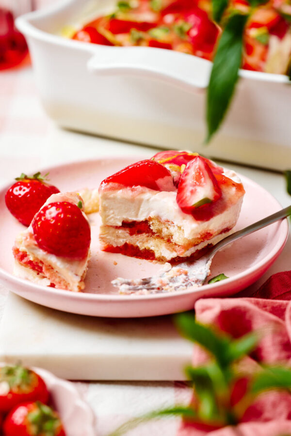 Eine Portion Erdbeer-Tiramisu auf einem Teller mit einer Gabel, einem Schälchen mit frischen Erdbeeren und Minzblättchen im Vordergrund und eine Keramikform mit dem Tiramisu im Hintergrund.