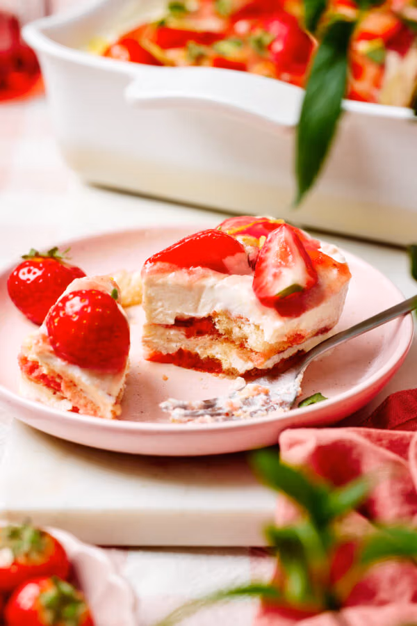 Eine Portion Erdbeer-Tiramisu auf einem Teller mit einer Gabel, einem Schälchen mit frischen Erdbeeren und Minzblättchen im Vordergrund und eine Keramikform mit dem Tiramisu im Hintergrund.