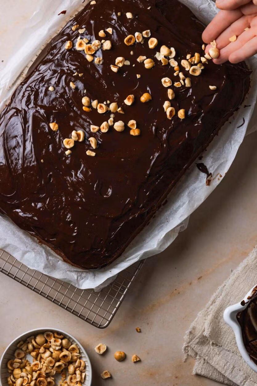 Geröstete Haselnüsse werden auf die Schokoladen-Glasur auf einen Zucchinikuchen vom Blech gestreut.