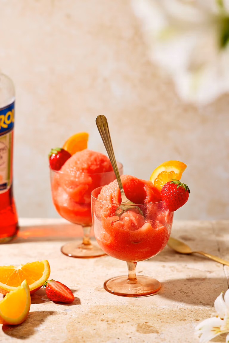 Frozen Aperol Spritz in Cocktailgläsern mit goldenen Löffeln, garniert mit Erdbeeren und Orangen, im Sonnenlicht.