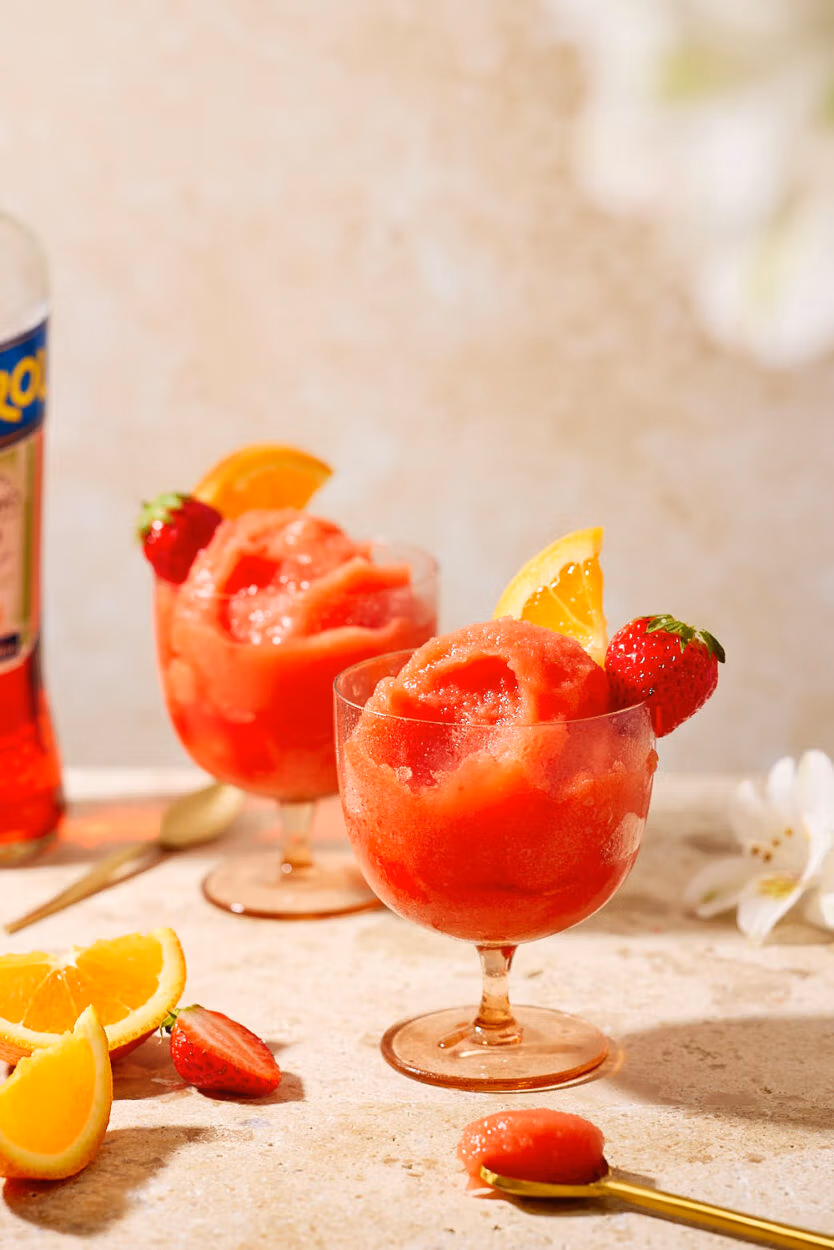 Nahaufnahme zweier Gläser mit Frozen Aperol Spritz, Erdbeeren und Orangen, auf heller Steinplatte.