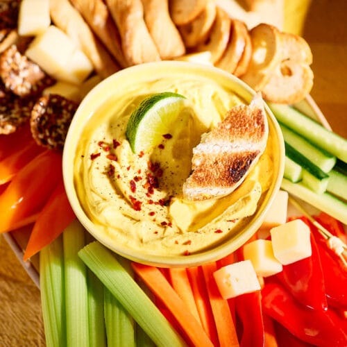 Nahaufnahme eines cremigen Mango-Curry-Dips in einer gelben Schale, garniert mit Limettenscheibe und geröstetem Brot, serviert mit Gemüsesticks, Käsewürfeln und Crackern.