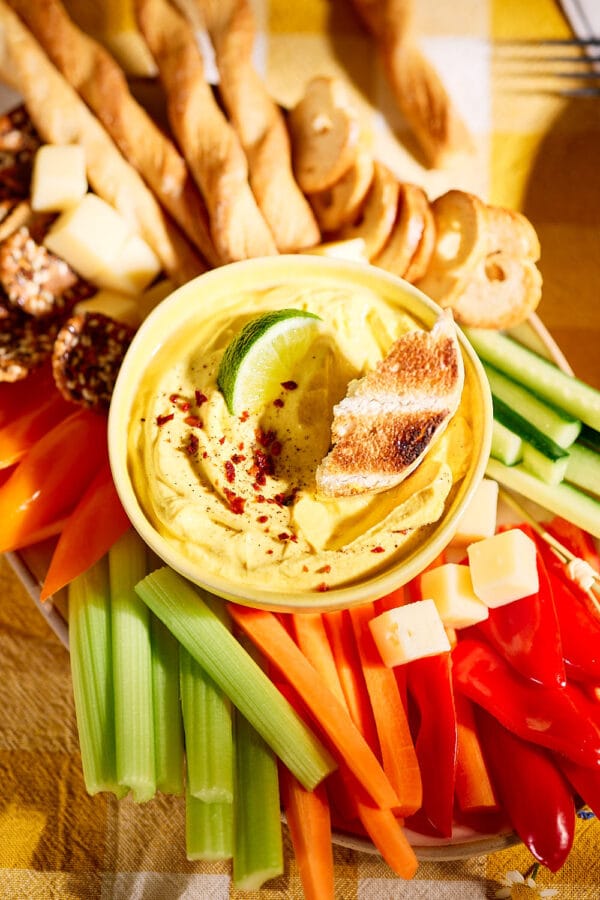 Nahaufnahme eines cremigen Mango-Curry-Dips in einer gelben Schale, garniert mit Limettenscheibe und geröstetem Brot, serviert mit Gemüsesticks, Käsewürfeln und Crackern.