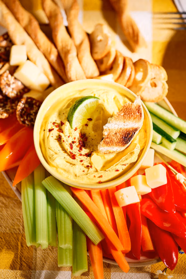 Nahaufnahme eines cremigen Mango-Curry-Dips in einer gelben Schale, garniert mit Limettenscheibe und geröstetem Brot, serviert mit Gemüsesticks, Käsewürfeln und Crackern.