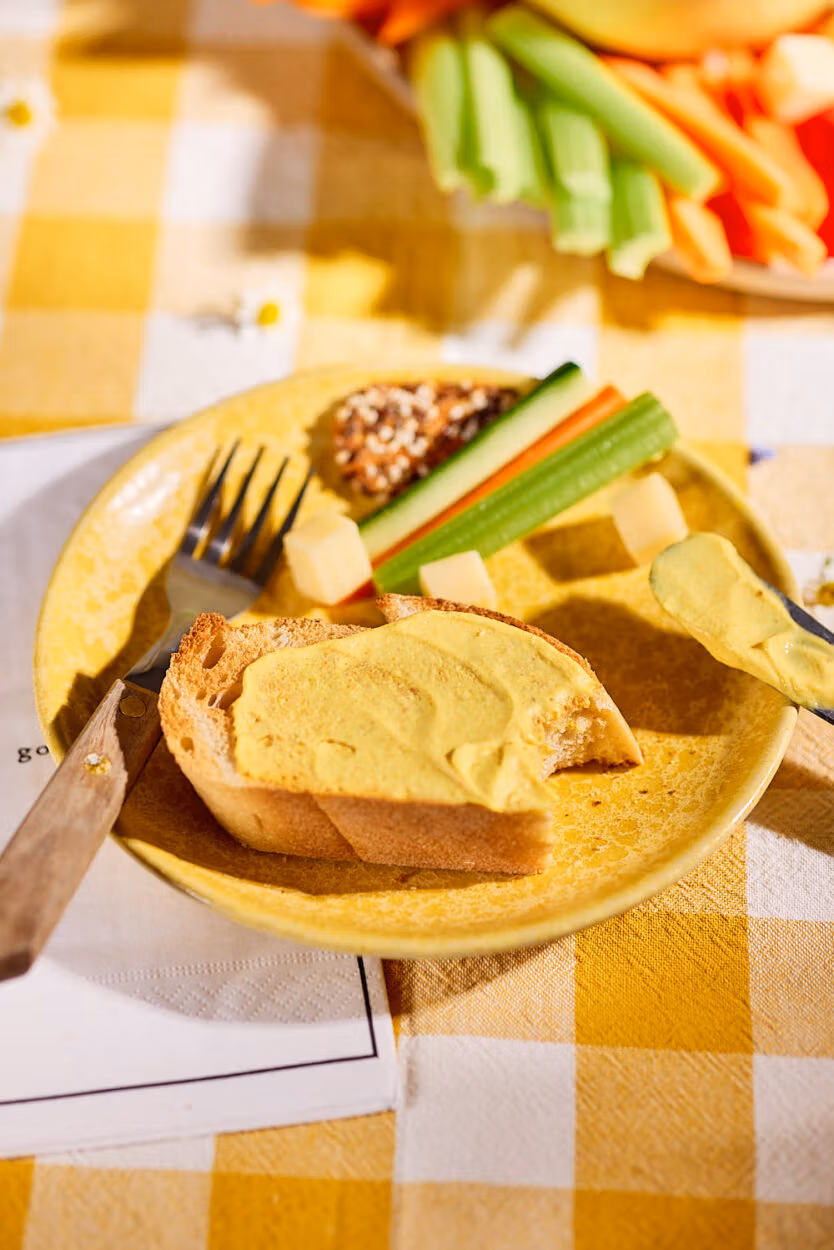 Brotscheibe mit Mango-Curry-Dip bestrichen, daneben frisches Gemüse und Käse auf einem gelben Teller bei sonnigem Picknick.