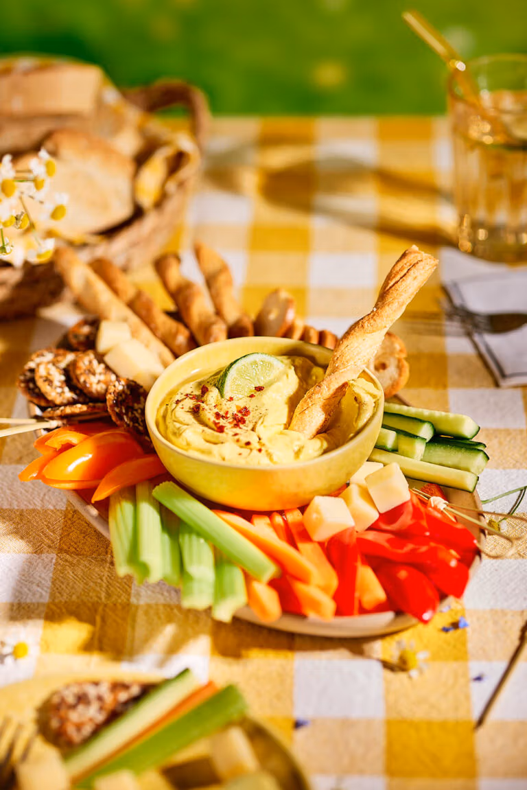 Gelber Mango-Curry-Dip mit Brotstücken in einer Schale auf einem bunten Vorspeisenbrett mit knackigem Gemüse, Käse und Grissini.