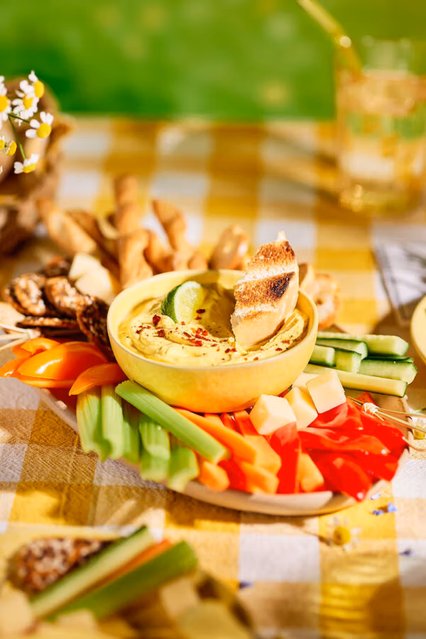 Bunter Picknicktisch mit Mango-Curry-Dip, Gemüsesticks, Käsewürfeln und Knabbergebäck, serviert auf kariertem Tuch und mit Blümchen dekoriert.