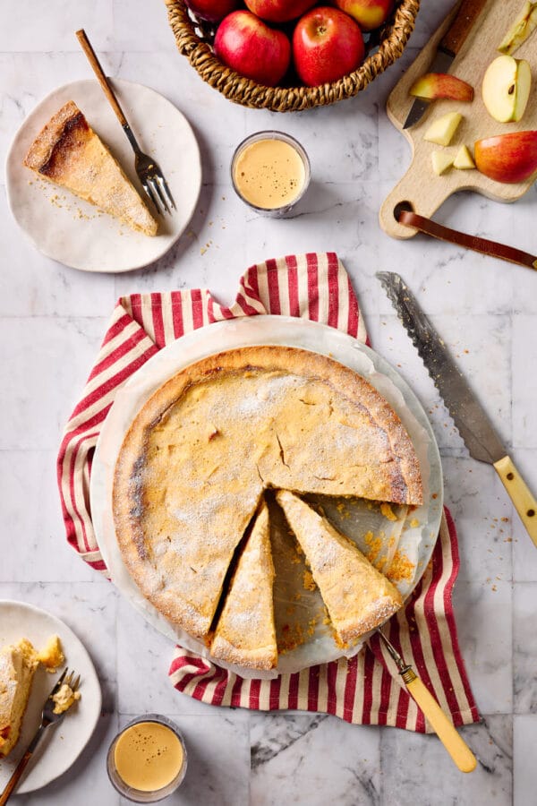 Ein angeschnittener Apfel-Quark-Kuchen mit zartem Mürbeteigboden in der Draufsicht, mit Äpfeln und Kuchentellern mit Kuchenstücken und Kaffeebechern drumherum.