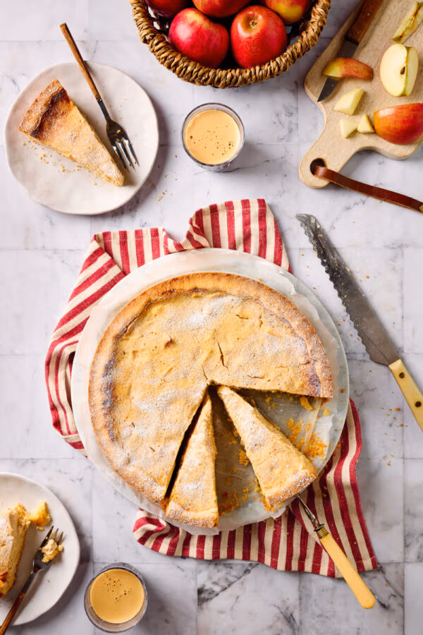 Ein angeschnittener Apfel-Quark-Kuchen mit zartem Mürbeteigboden in der Draufsicht, mit Äpfeln und Kuchentellern mit Kuchenstücken und Kaffeebechern drumherum.