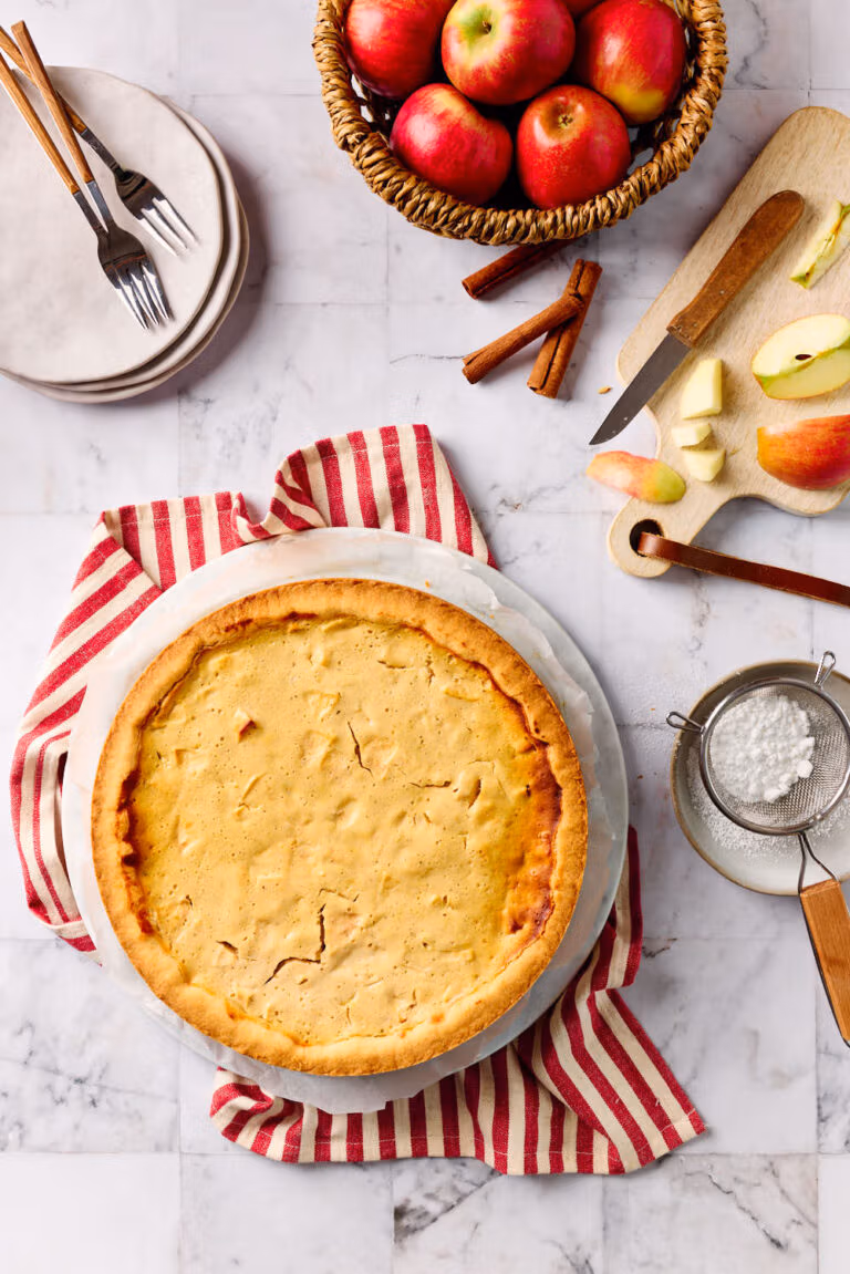 Saftiger Apfel-Quark-Kuchen mit zartem Mürbeteigboden in der Draufsicht, mit Äpfeln im Hintergrund und einem Sieb mit Puderzucker.