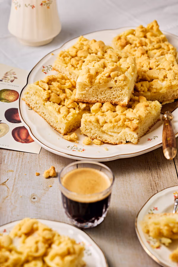 Apfelkuchen mit Hefeteig und Streusel auf einer Servierplatte mit Kuchentellern und einem Kaffee im Vordergrund.