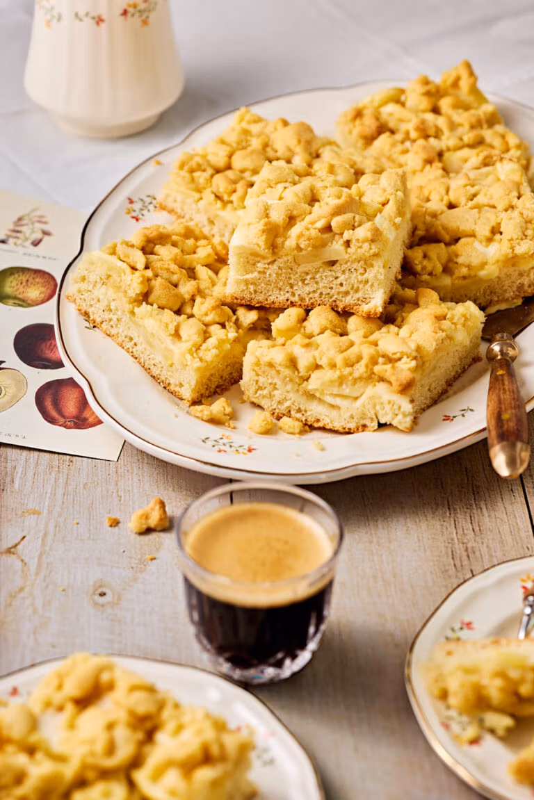 Apfelkuchen mit Hefeteig und Streusel auf einer Servierplatte mit Kuchentellern und einem Kaffee im Vordergrund.