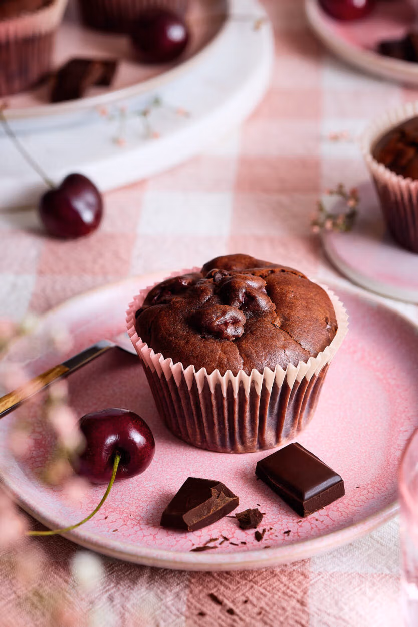 Nahaufnahme eines saftigen Schoko-Kirsch-Muffins mit gut sichtbaren Kirschstücken, daneben Schokolade und frische Kirschen auf einem rosa Teller.