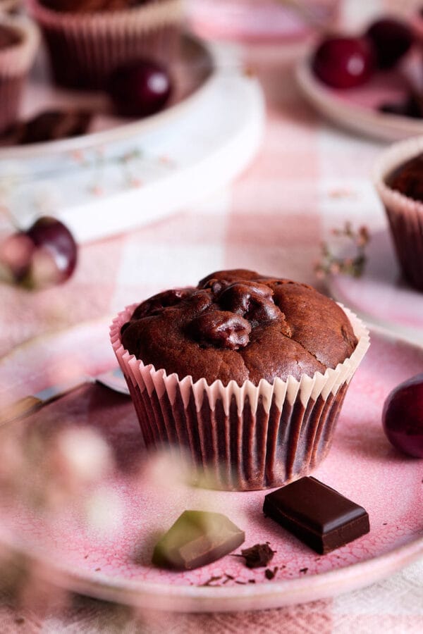 Detailaufnahme eines Schoko-Kirsch-Muffins mit glänzender Oberfläche, frischen Kirschen und dunkler Schokolade auf rosafarbenem Geschirr.