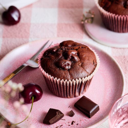 Stimmungsvolle Szene mit einem Muffin auf rosa Teller, Kirschen, Schokolade und zartem Blütendekor – sommerlich und einladend arrangiert.