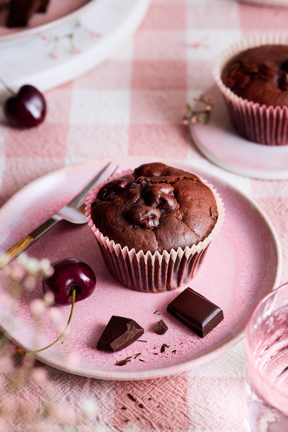 Stimmungsvolle Szene mit einem Muffin auf rosa Teller, Kirschen, Schokolade und zartem Blütendekor – sommerlich und einladend arrangiert.