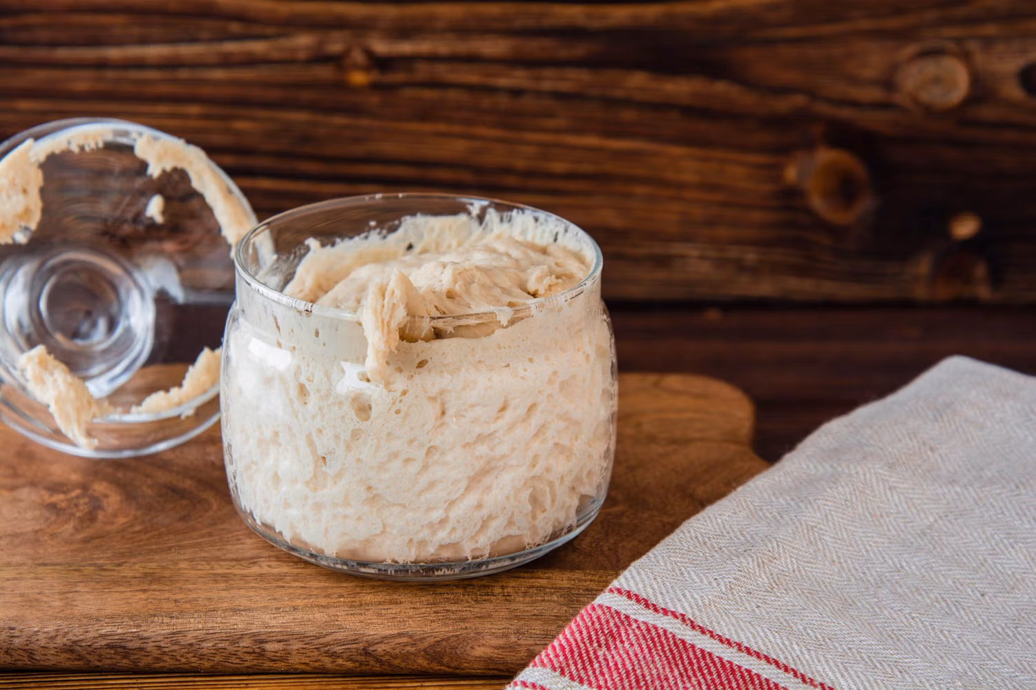 Reifer Lievito Madre aus Hefewasser in einem Glas auf einem Holzbrett mit einem roten Geschirrtuch daneben.