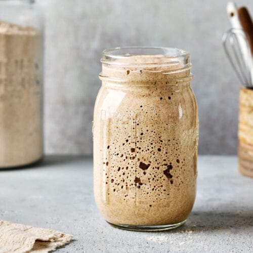 Frisch gefüttert und reif: Sauerteig in einem Glas mit einem Mehlglas im Hintergrund.