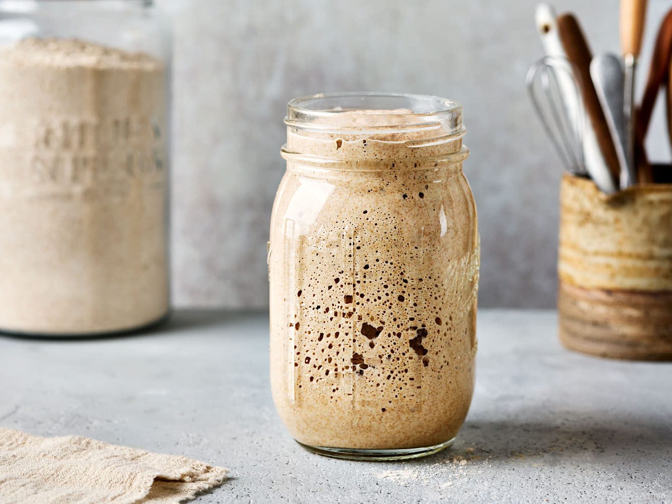 Frisch gefüttert und reif: Sauerteig in einem Glas mit einem Mehlglas im Hintergrund.