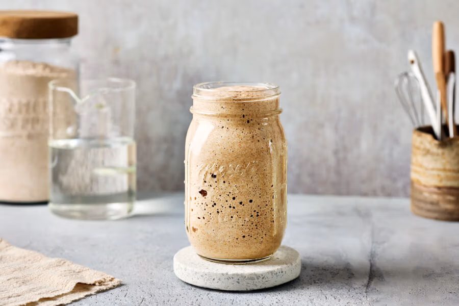 Reifer Dinkelsauerteig in einem Glas mit einem Glas Mehl und einer Karaffe mit Wasser sowie Löffel und Schneebesen im Hintegrund.