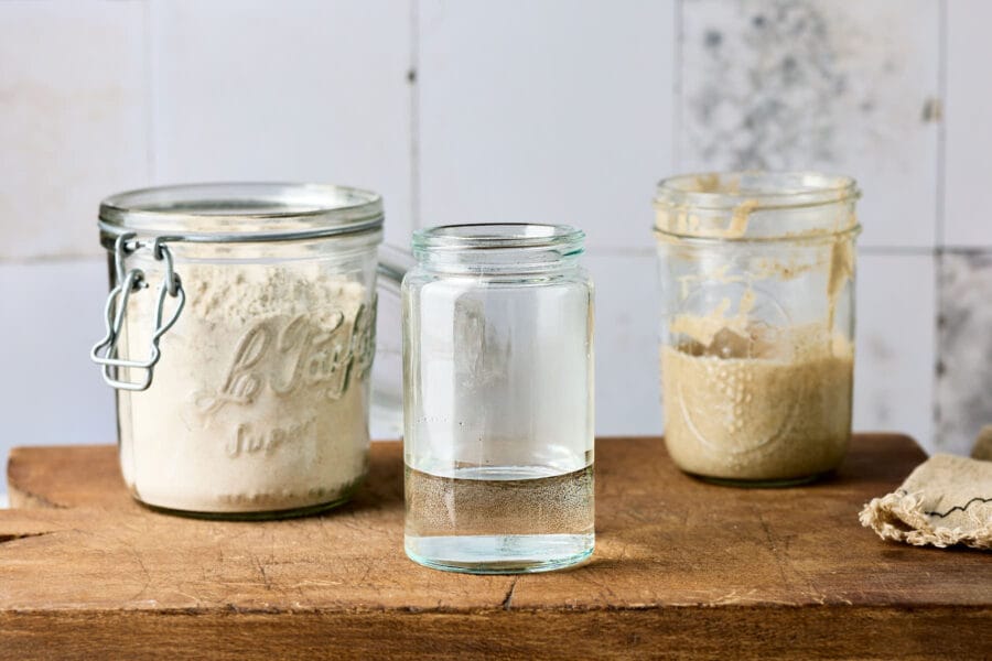 Wasser in einem Einmachglas zum Sauerteigfüttern, mit Mehl und Sauerteiganstellgut im Hintergrund.